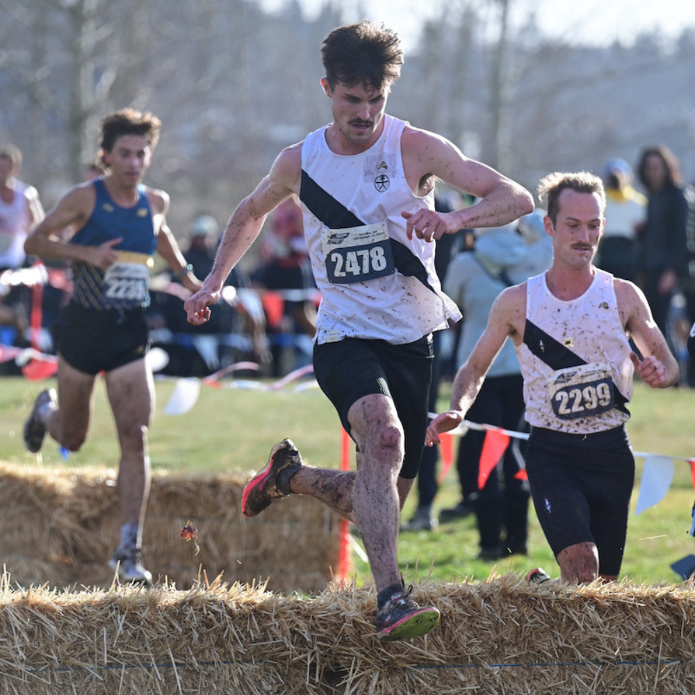 Male runner wearing gray SockMoose running socks in a race jumping over a hay bail